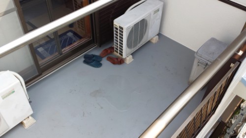 Balcony scene with an outdoor A/C unit on the floor, two pairs of blue and brown slippers, and a gray cooler near the railing.
