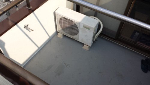 Outdoor air conditioning condenser unit mounted on a balcony floor, with railing and a pair of slippers in the corner.