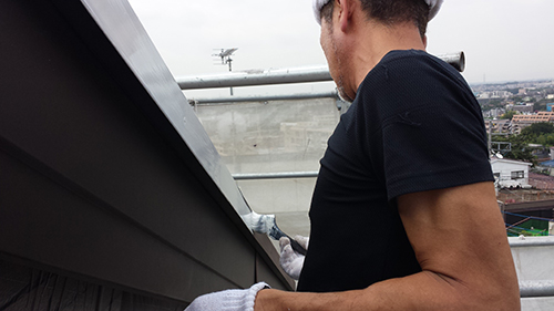 koushin_03hafu2 | 外壁塗装は神奈川県横浜市の塗装職人 Man on a rooftop wearing a black T-shirt and gloves, working along the edge of a building with a cityscape in the background.