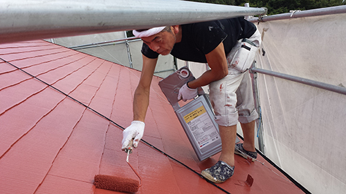 koushin_04uwanuri | 外壁塗装は神奈川県横浜市の塗装職人 Worker on a sloped red roof applying paint with a roller, paint can in hand.