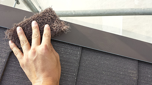 koushin_05keren1 | 外壁塗装は神奈川県横浜市の塗装職人 A hand presses a clump of brown insulation fibers onto a roof seam above dark shingles with scaffolding in the background.