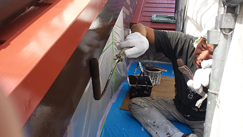 koushin_07hafu1 | 外壁塗装は神奈川県横浜市の塗装職人 Tradesperson on a scaffold painting a metal beam with orange-red paint using a roller; blue tarp on the ground and a paint bucket nearby.