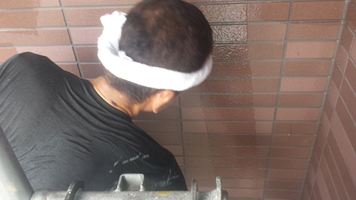 koushin_09gaiheki2 | 外壁塗装は神奈川県横浜市の塗装職人 Back view of a person wearing a white cloth headband and a black shirt, standing against a brown brick wall in a narrow space.