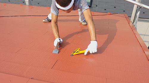 koushin_10engiri2 | 外壁塗装は神奈川県横浜市の塗装職人 Worker wearing gloves trims red roofing material on a sloped roof with a utility knife and scraper nearby