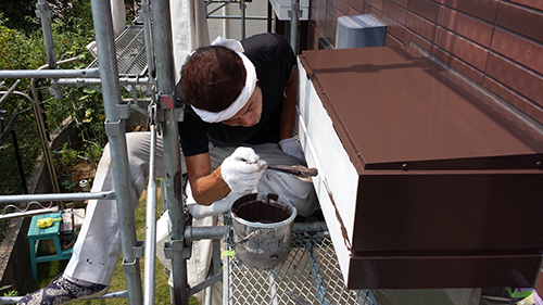 koushin_12hafu1 | 外壁塗装は神奈川県横浜市の塗装職人 Construction worker on scaffolding painting a brown metal vent box on the exterior wall with a brush.