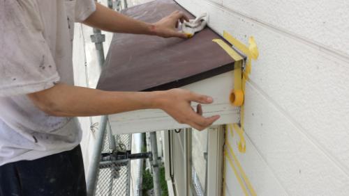 Construction worker attaching a brown metal panel to a white exterior wall, taping edges with yellow painter's tape while standing on scaffolding.