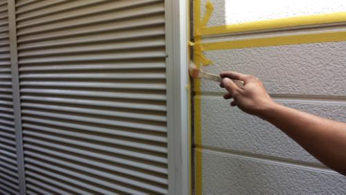 Person applies yellow painter's tape along the seam between a vent and a painted exterior wall to prep for painting
