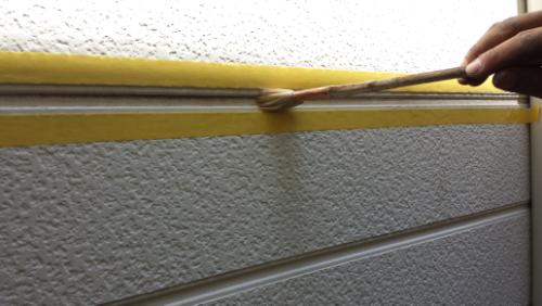 Person applying sealant along a seam on a textured exterior wall using a small brush; yellow masking tape is visible on the edge.