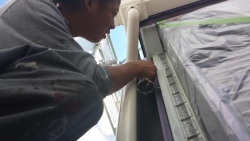 Worker in a gray shirt paints a window frame from the outside, with plastic sheeting masking the area around the window.