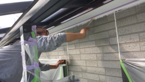 Person paints an exterior brick wall under a balcony, with masking tape and plastic sheeting protecting nearby surfaces.