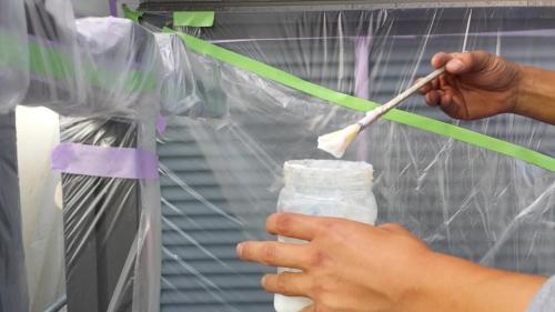 Hands brush white material onto a jar inside a plastic-covered workspace.