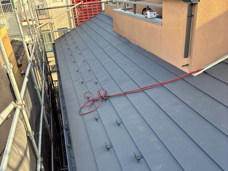 260422_14 | 外壁塗装は神奈川県横浜市の塗装職人 Overhead view of a gray metal sloped roof under construction, with scaffolding along the left edge and a red extension cord looped across the surface.