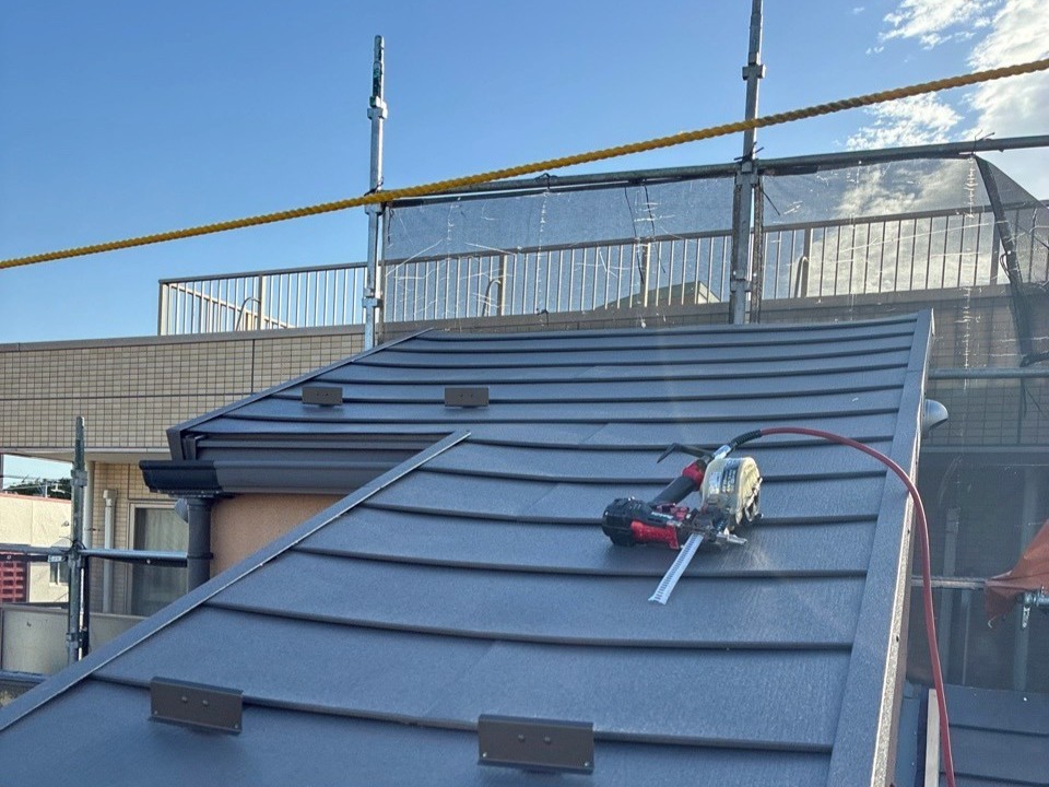 L260422_11 | 外壁塗装は神奈川県横浜市の塗装職人 Roof under construction with metal tile panels; cordless drill and tape measure sit on the slope, scaffolding and safety netting in the background.