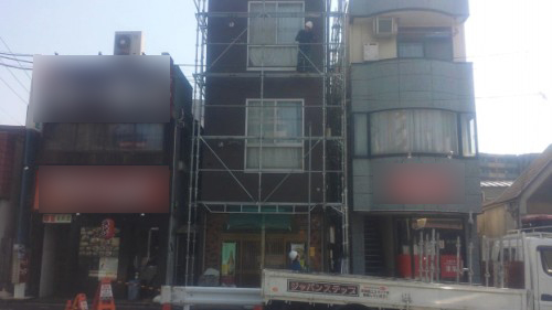 Three-story building under renovation with metal scaffolding, a worker on the scaffold, and a truck parked on the street.”] ,